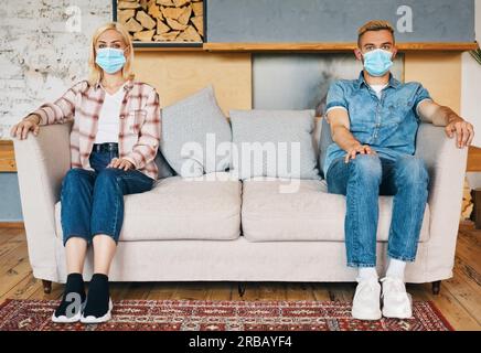 Man and woman in facemask sitting on sofa at home. Social distancing, coronavirus concept Stock Photo
