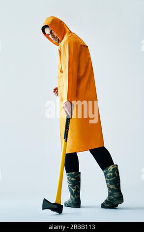 Full length portrait of young spooky man in bright raincoat holding big axe on white background Stock Photo