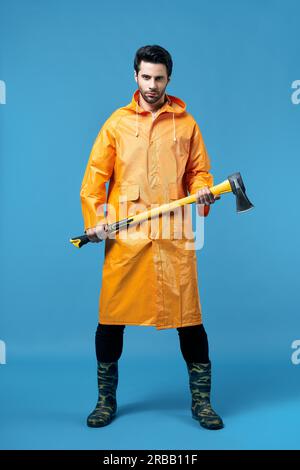Full length portrait of young handsome man in bright raincoat holding big axe. Brutal male, lumberjack, fisherman with ax Stock Photo
