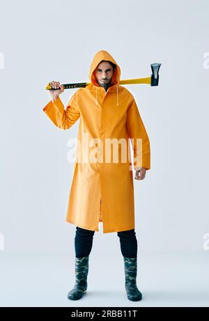 Full length portrait of young handsome man in bright raincoat holding big axe. Brutal male, lumberjack, fisherman with ax Stock Photo