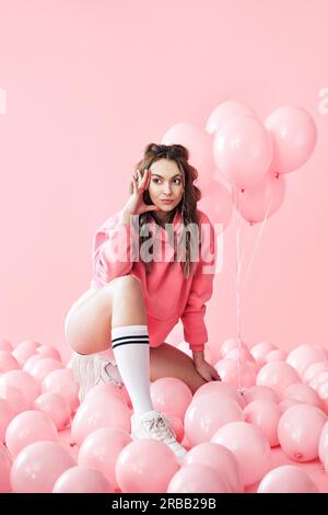 Bored fashionable woman sitting on floor with pink air balloons on pink pastel background. Celebration, bad party, disappointment concept Stock Photo