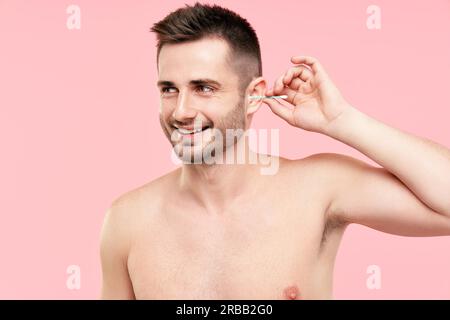 Young smiling man cleaning ear with cotton swab over pink background. Hygiene concept Stock Photo