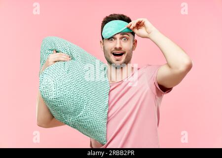 Funny cheerful man in sleeping mask holding pillow in hands looking to camera over pink background. Relax, good morning concept Stock Photo
