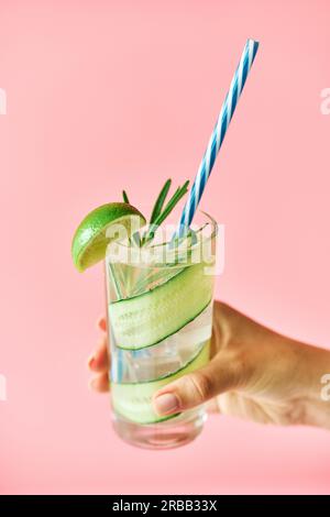 Young woman hand holding cold refreshing cocktail with lime, cucumber, rosemary and ice in glass on pink background close up. Party, summer concept Stock Photo