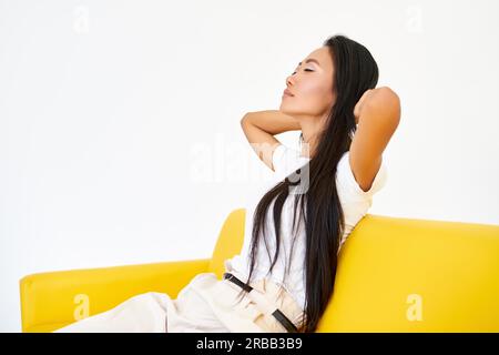 Young pretty woman closed her eyes sitting on yellow sofa and relaxing after busy day. Rest, comfort concept Stock Photo