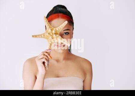 Pretty young afro woman covering her eye with starfish. Female beauty, spa, clean skin concept. Copy space Stock Photo