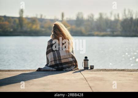 Woman relaxing alone by a river in the city wrapped in wool blanket enjoying nature and drinks tea from thermos. Picnic, rest, travel concept. Back Stock Photo