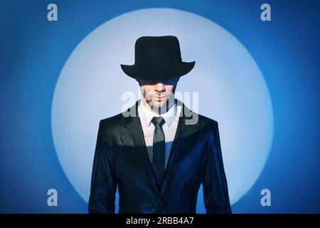 Portrait of handsome man in black coat hiding face behind his hat in the spotlight on studio background. noir film style. Private detective Stock Photo