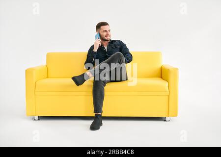 Smiling relaxed man talking on the phone while sitting on yellow sofa over white background Stock Photo
