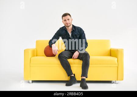 Young trendy man sitting with basketball ball on yellow sofa over white background. Sport concept Stock Photo