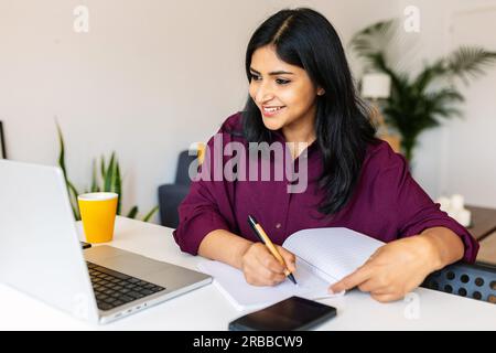 Young adult indian student woman taking notes while using laptop at home Stock Photo