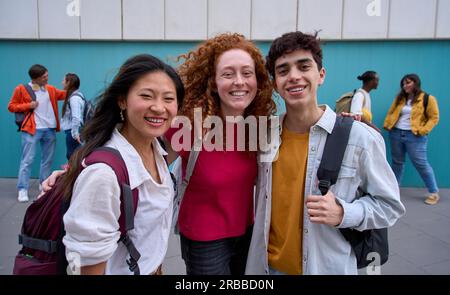 Group portrait of three smiling multiracial students hugging with backpacks looking at camera. Stock Photo