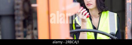 Young asian woman is foreman using radio for communication while driving forklift at distribution warehouse, engineer working and shipping container a Stock Photo