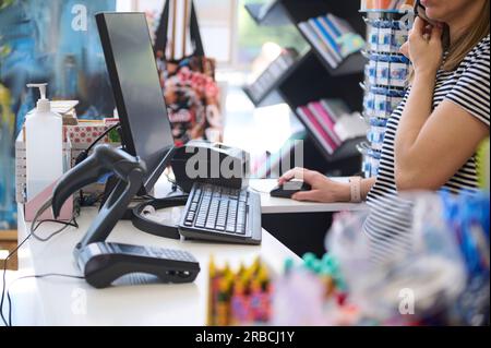 Woman seller in office supply shop, talks on mobile phone, works on PC computer, orders merchandise, standing at countertop with retail barcode scanne Stock Photo