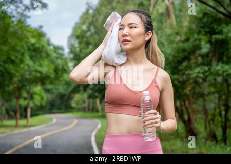 Young attractive Asian woman wiping off her sweat from her forehead during her water break from her morning exercise run at a running track of a local Stock Photo