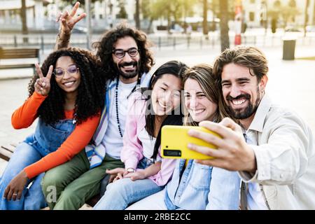 Diverse group of friends taking a selfie in the street sitting on a bench. Cheerful multiracial group of young hipsters taking a picture outside in a Stock Photo