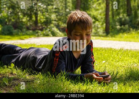 A boy is lying in the park on the grass with a phone and laughing. Stock Photo
