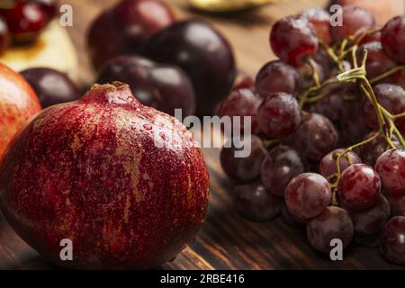 A ripe pomegranate with water drops surrounded by red grapes and plums Stock Photo