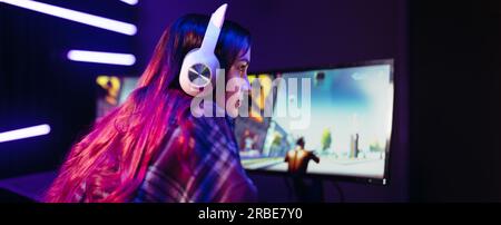 Gamer concentrating on her video game as she sits in front of a multi-monitor setup with a headset. Young woman getting immersed in the realm of digit Stock Photo