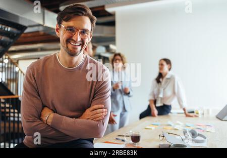 Successful business man sitting in a boardroom with his team in the background. Happy business professional looking at the camera in a meeting. Busine Stock Photo
