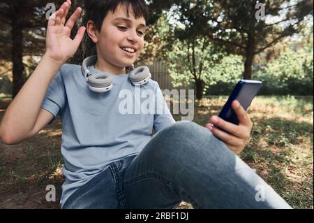 Toned portrait. Teen school boy sitting under a tree in a park, greeting his friend while talking on mobile phone via video link. Online conference. I Stock Photo