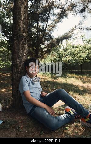 Toned full length portrait of multi-ethnic cheerful teenager, handsome schoolboy sitting under a tree during a recreation after class, smiling looking Stock Photo