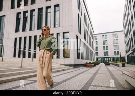 Smiling woman office worker is talking by phone while going on modern building background  Stock Photo