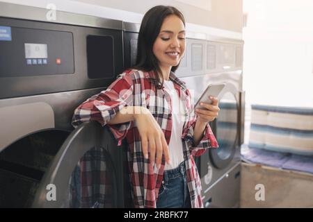 Happy Young Lady Using Cellphone While Doing Laundry Indoor Stock Photo