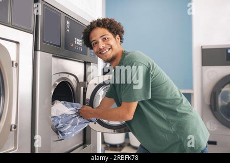 Latin Young Guy Loading Washing Machine Posing At Laundrette Room Stock Photo