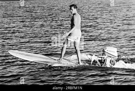 Dana Point, California:  c. 1960 Surfing champion and innovator Hobie Alter demonstrates the ultimate in surfboards with a 5 horsepower Johnson outboard motor attached to his surfboard. Stock Photo