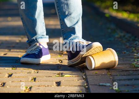 a man's foot steps on a paper cup lying on a path in the park. man littering in the park. environmental pollution. garbage in the city Stock Photo