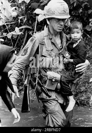 An Khe, Vietnam:  1967 A member of the 1st Calvary Division helps a Vietnamese woman and her child across a stream. Stock Photo