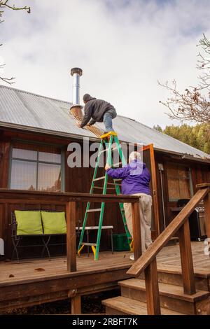 Father holding ladder for son to craw up on tin roof of cabin in winter to work on stove pipe Stock Photo