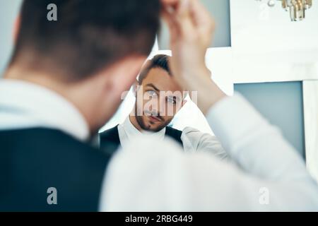 Handsome young man touching his hair with hand and smiling while standing in front of the mirror Stock Photo