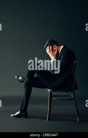 Sad unhappy man in black suit sitting on the chair and closed face by two hands. Profile view portrait. Depression and pain concept Stock Photo