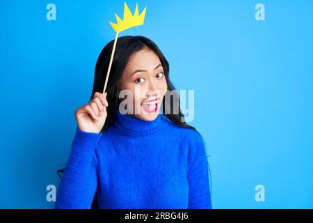 Emotional suprised woman with paper crown on stick on blue background. Party, emotions concept Stock Photo