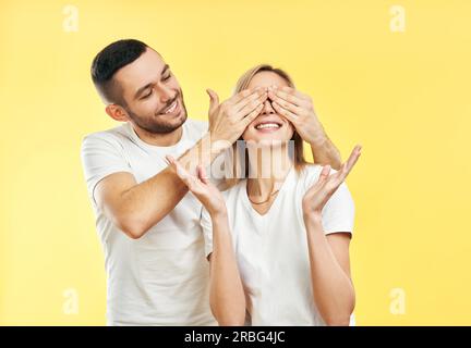 Guess who. Surprise for her. Young man covering eyes of his girlfriend isolated on yellow background Stock Photo