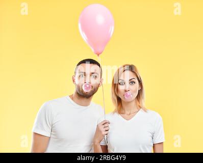 Young couple blowing bubble with chewing gum and holding pink air balloon on yellow background Stock Photo