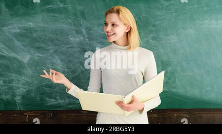 Education in high school university college. Back to school. World teachers day. Smiling female teacher with notebook standing near blackboard giving Stock Photo