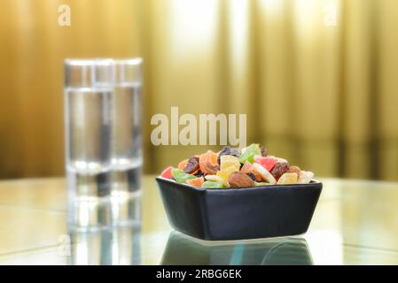 Many colorful dry fruits in a little black square bowl put on a glass table, with the reflection of two vodka glasses Stock Photo