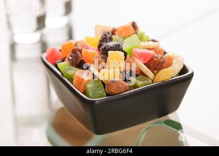 Many colorful dry fruits in a little black square bowl put on a glass table, with the reflection of two vodka glasses Stock Photo