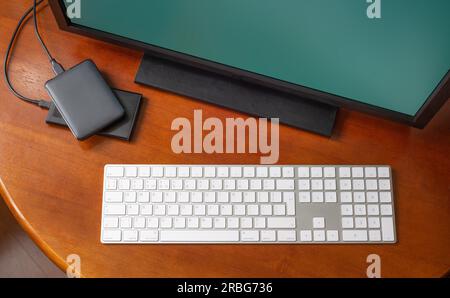 A computer monitor, a digital wireless keyboard and hard disk drives on a round wooden desk Stock Photo