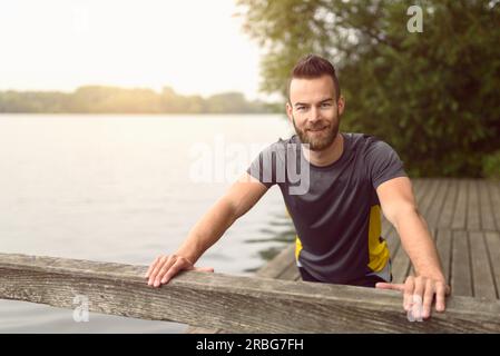 Young man doing stretching exercises on a wooden deck overlooking a lake as he smiles at the camera in a healthy lifestyle concept Stock Photo