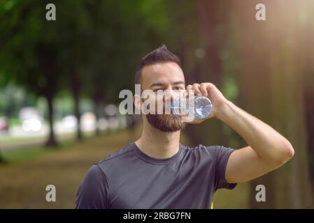 Young man with a beard drinking bottled water as he walks down a tree lined avenue in a park, close up head and shoulders Stock Photo