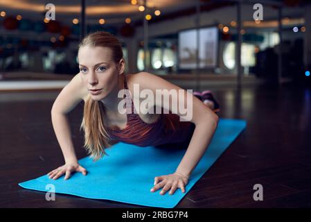 Young blonde woman doing push-up exercise in gym and looking at camera while lying on blue yoga carpet over mirror background Stock Photo