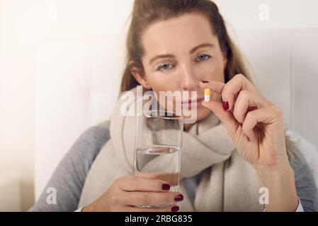 Portrait of a sick woman in bed looking at camera upset while holding a glass of water and a pill against flu symptoms Stock Photo