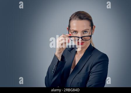 Stylish business executive peering over her glasses at the camera with a thoughtful serious expression on a grey graduated background Stock Photo