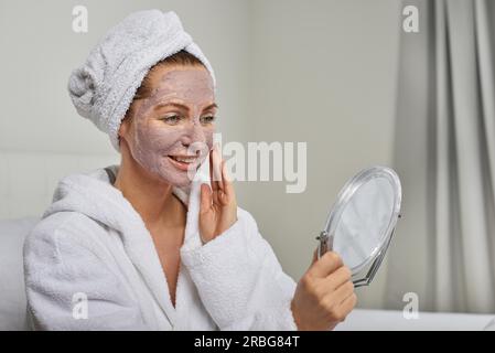 Attractive woman in a white towelling robe applying a face mask while looking in a handheld mirror as she pampers herself with a beauty treatment Stock Photo