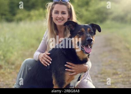 Pretty blond woman with her two dogs crouching with an arm around each at the side of a rural road with long spring grass and focus to a large brown Stock Photo