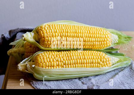 Close up of fresh corn cobs with leaves on a wooden table over grey wall background. Raw corncobs on light brown wooden table, rustic style. Corn on c Stock Photo